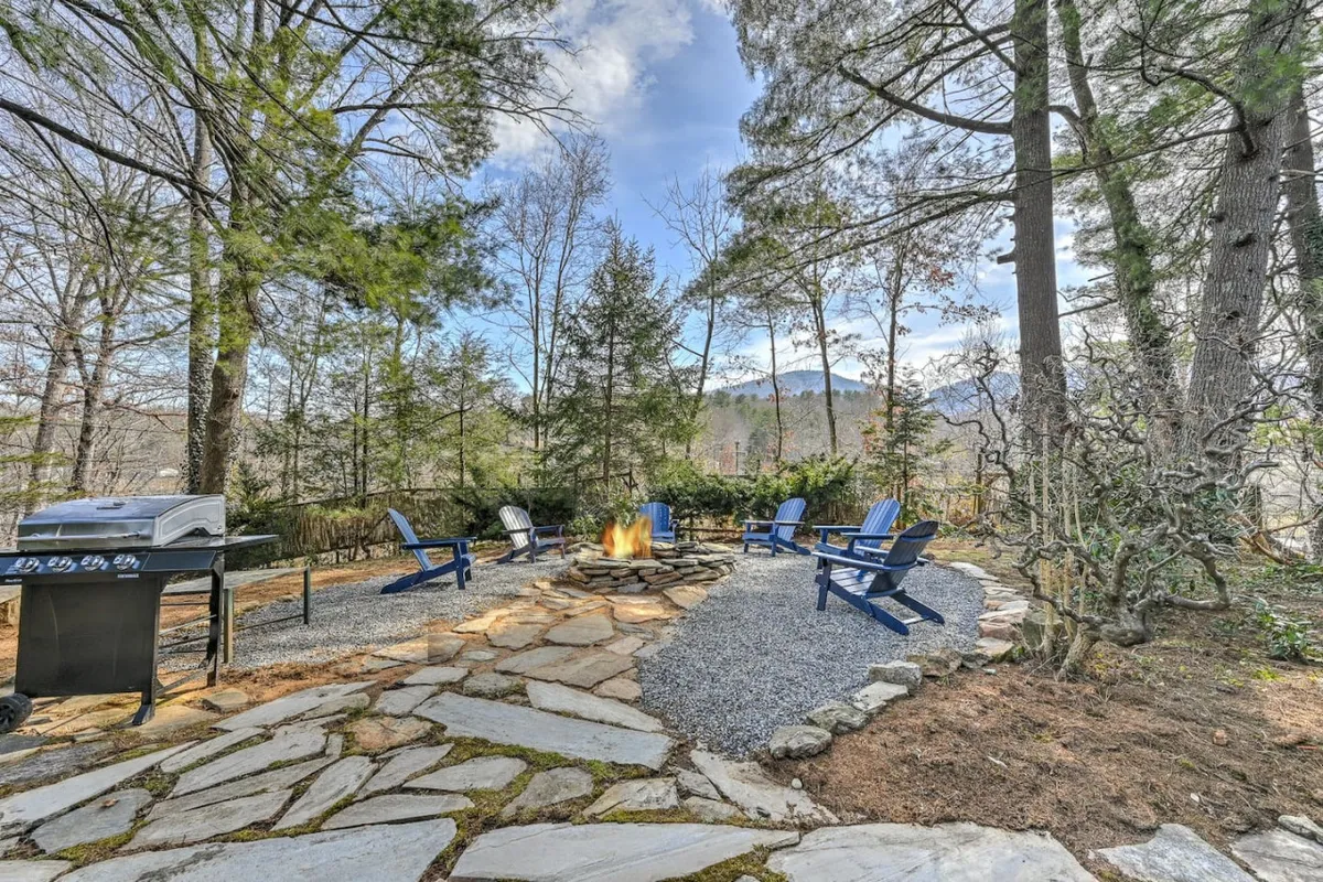 An inviting outdoor fire pit scene surrounded by Adirondack chairs and enveloped by trees. The fire pit takes center stage, creating a cozy, communal space ideal for group gatherings, storytelling, or roasting marshmallows under the stars. A grill is visible, suggesting the potential for outdoor meals and camaraderie.