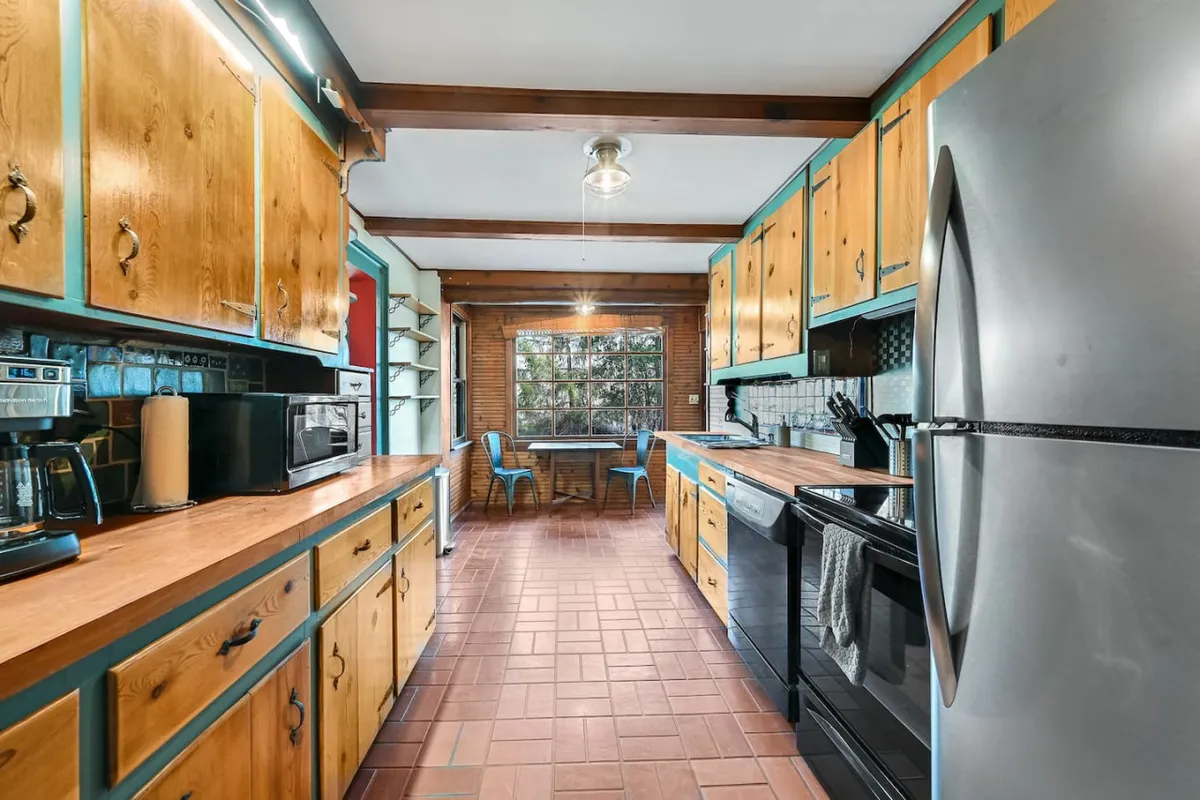 A kitchen and dining area with wooden cabinetry, stainless steel appliances, a dining area with blue chairs, and a large window that brings in natural light. The setup makes it easy to imagine preparing meals and sharing them in a welcoming setting.