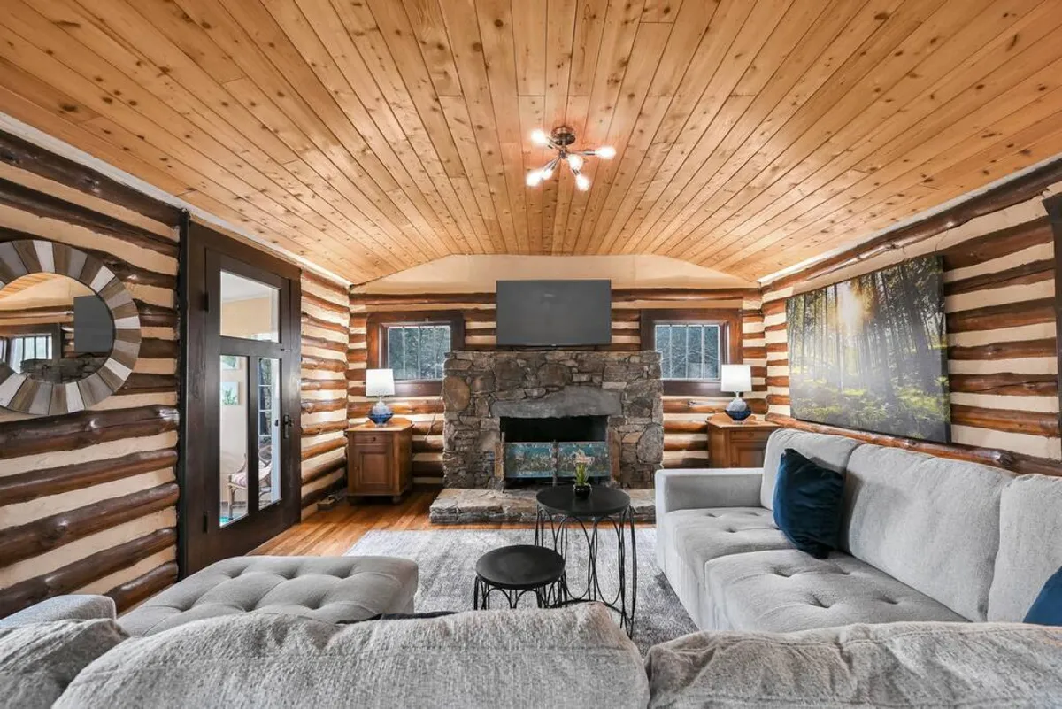 A spacious living room featuring a stone fireplace as the focal point, with a comfortable sectional sofa arranged around it. Large windows let in abundant natural light, and carefully chosen decorative artwork adds visual interest to the space, making it feel both inviting and stylish.