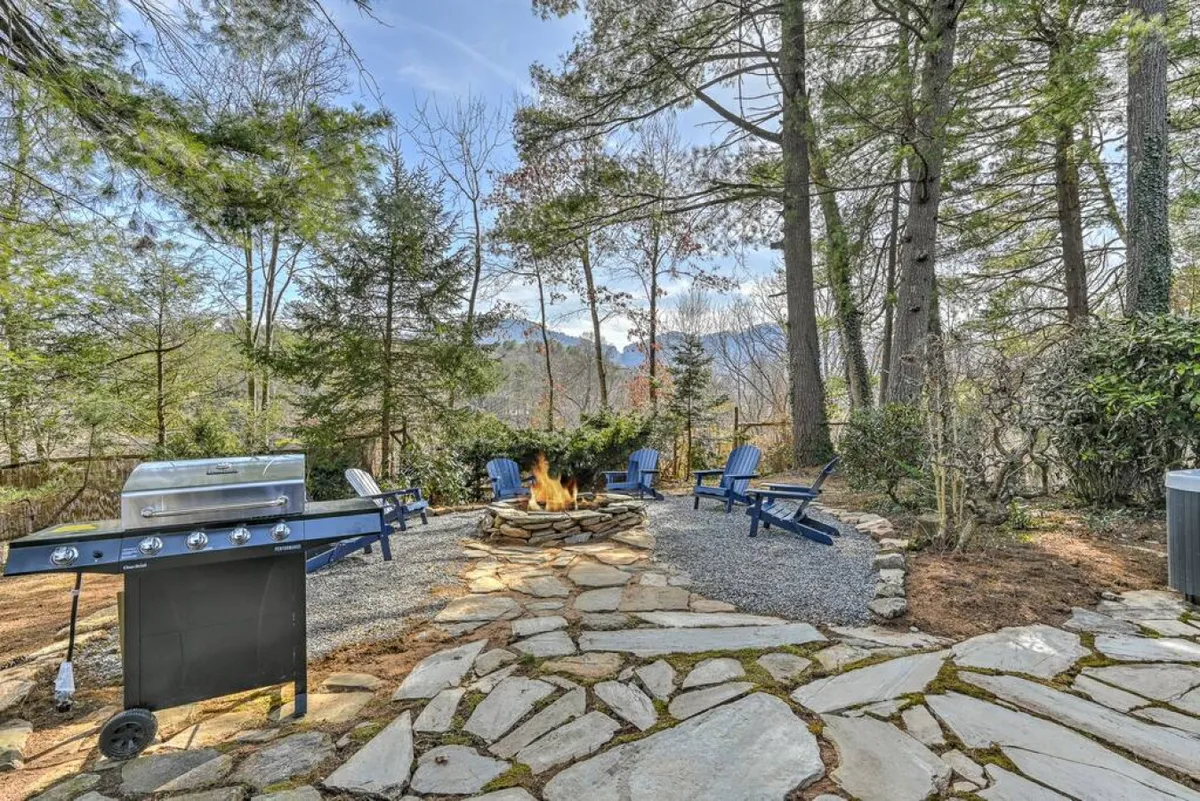 An inviting outdoor area featuring a large stone fire pit surrounded by Adirondack chairs, a grill nearby, and a natural stone pathway leading through a wooded backyard. The scene is set for family gatherings, campfire evenings, and outdoor barbecues, with plenty of space for everyone to relax and play.