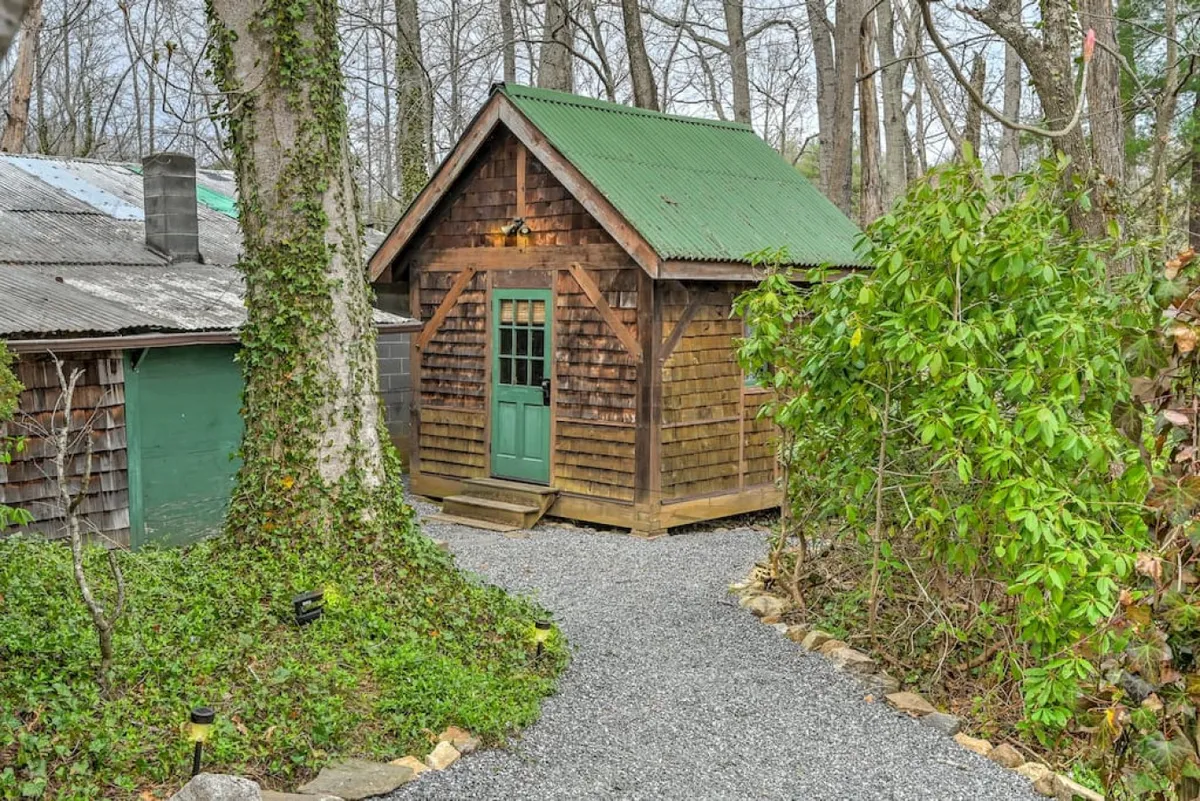 An exterior view of a quaint, stand-alone wooden cabin with a green door and surrounded by lush greenery and a gravel pathway. The cabin exudes privacy and peacefulness—ideal for those seeking a unique and secluded sleeping option.