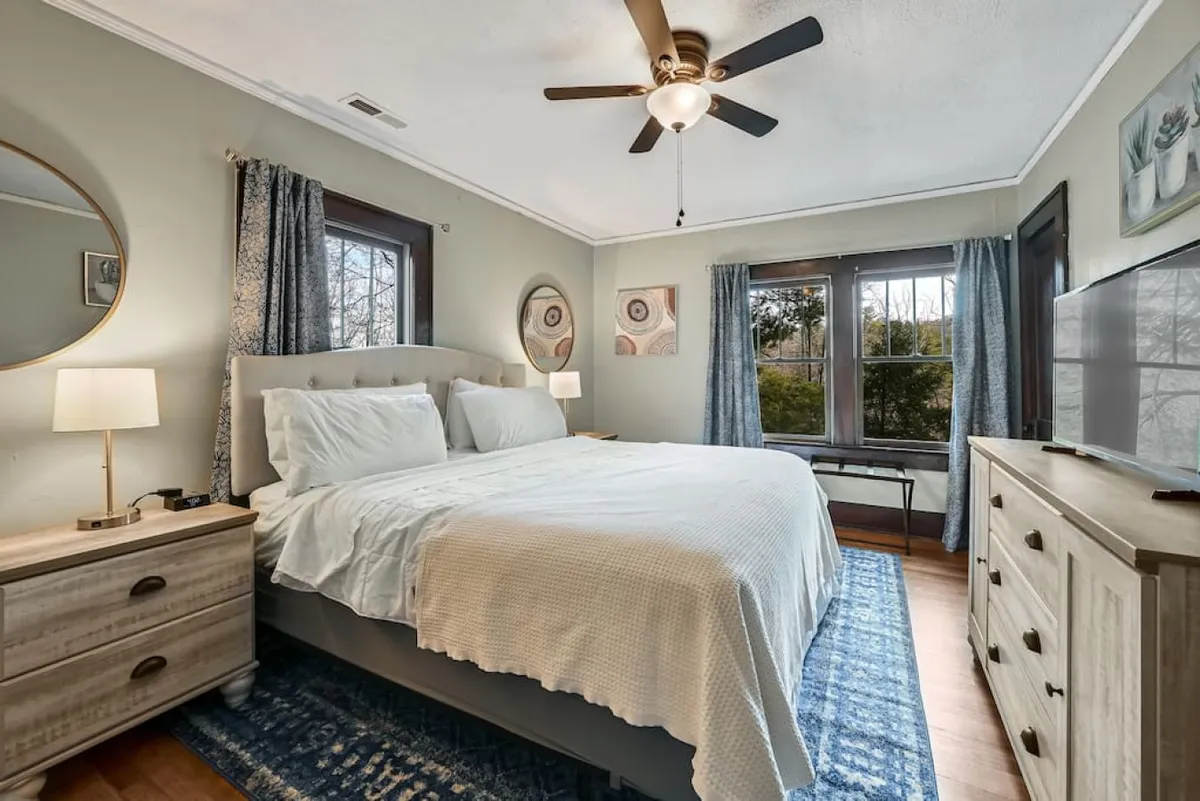 A serene bedroom with a king-size bed, dressed in crisp white linens and a decorative rug, accompanied by a nightstand with a lamp and a window offering natural light. The space feels tranquil and inviting, perfect for unwinding after a long day.