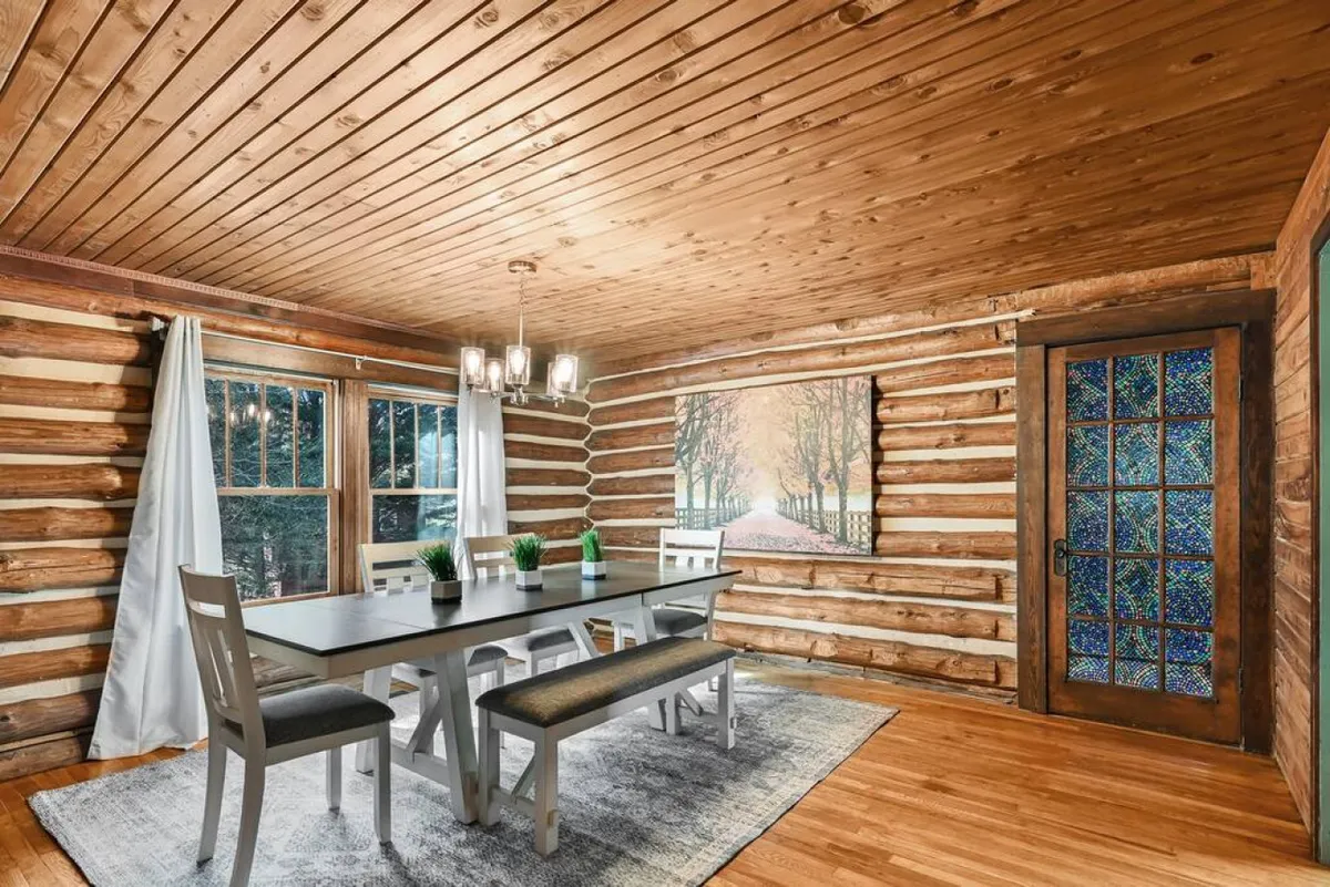 A warmly lit dining space featuring a wooden dining table surrounded by chairs, rustic wooden logs, and decorative artwork. A stained glass door adds character, inviting guests to gather for meals and celebrations.
