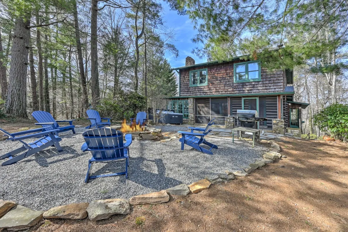 A cozy fire pit setup with blue Adirondack chairs on a stone patio, the house visible in the background. The scene invites gatherings, storytelling, and evening relaxation, with a communal, welcoming vibe.