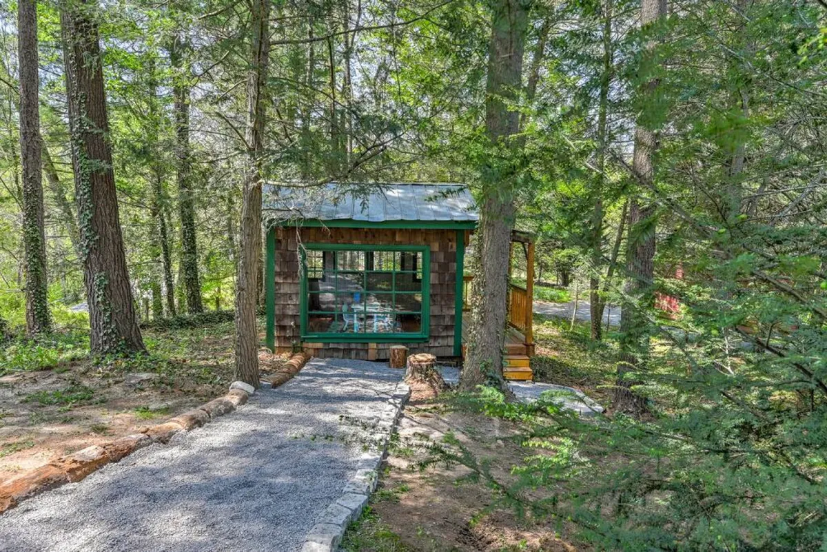 A small, quaint cabin nestled among trees, with a gentle pathway leading up to it. The setting is serene and private, hinting at opportunities for guests to retreat, read, or enjoy solitary moments amidst nature, away from the main communal spaces.