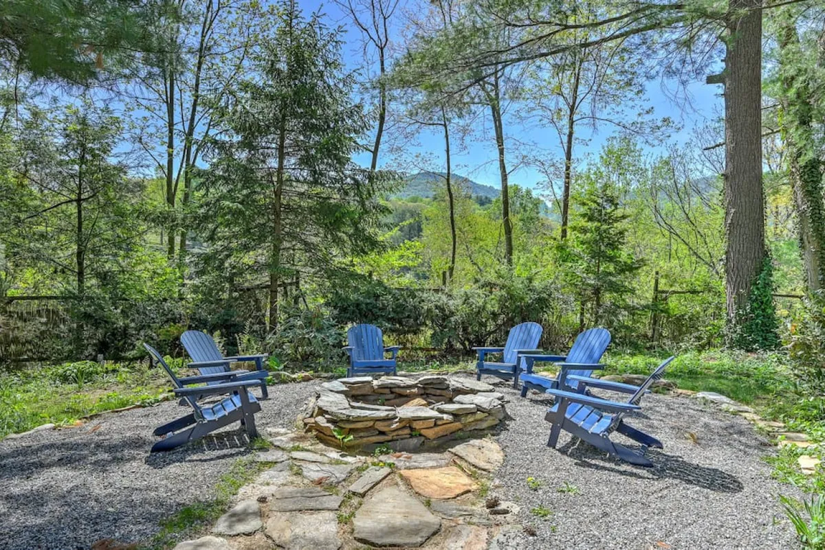 This image showcases a cozy outdoor fire pit area with blue chairs arranged in a circle, surrounded by lush trees. The setting is ideal for group gatherings, storytelling, and relaxing in nature, with ample open space that could easily accommodate pets at play.