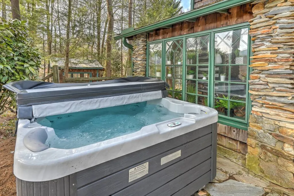 An outdoor area featuring a bubbling hot tub nestled beside a stone wall, with green-trimmed windows and lush greenery in the background. The space is tranquil and designed for relaxation and socializing, perfect for stargazing or enjoying the mountain air.
