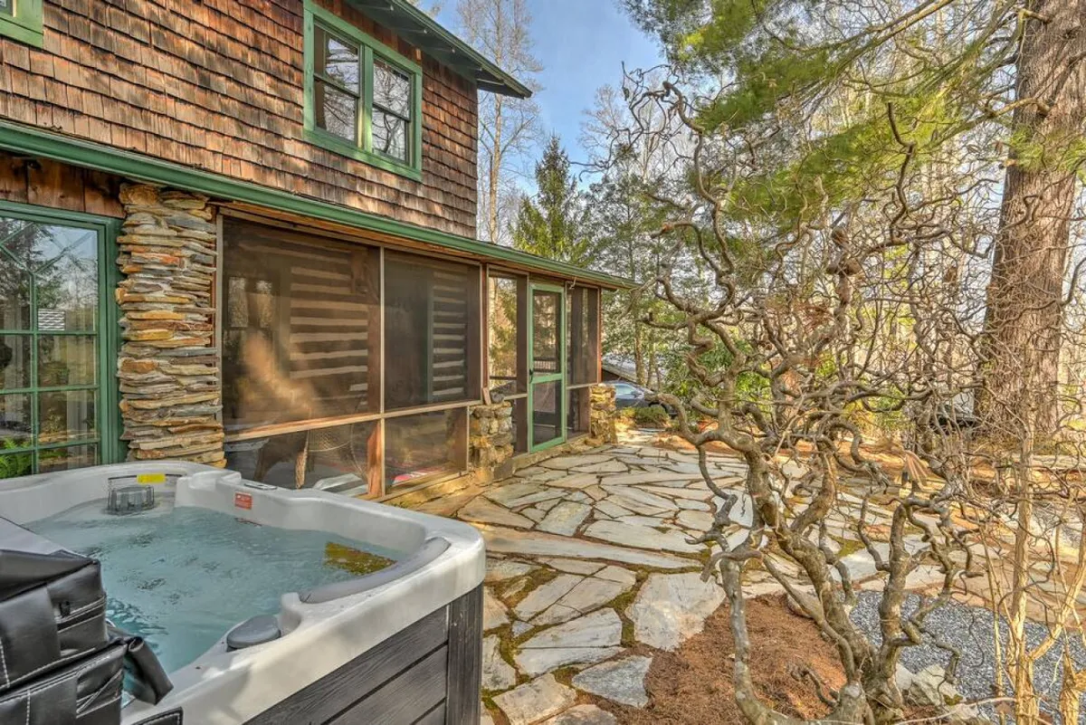 A tranquil exterior patio scene featuring a hot tub set along a natural stone pathway, adjacent to the wooden house. Surrounded by nature, the hot tub offers a relaxing spot for guests to unwind after a day of games and activities, with plenty of space around for socializing or quiet contemplation.