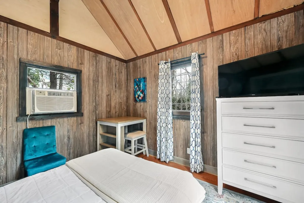 A peaceful bedroom scene featuring a bed with white bedding, a blue accent chair, a wooden table, a television, and a window with curtains letting in soft light. The decor feels modern yet warm, ideal for relaxing or enjoying a quiet evening in.