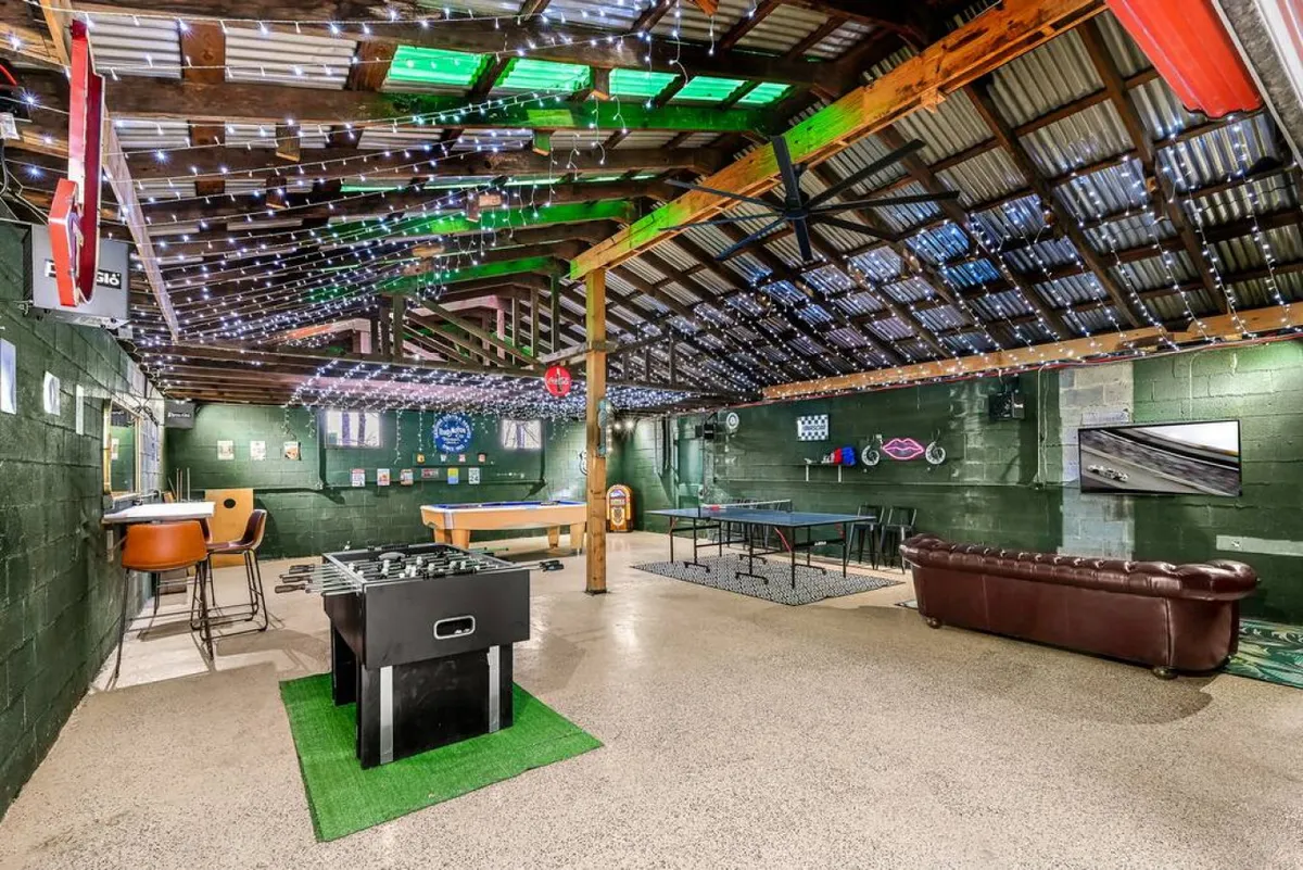A welcoming view of the game room featuring a foosball table, pool table, ping pong table, and a chocolate brown leather couch. The bar area with stools completes the scene, inviting guests to relax, play, and socialize. The room exudes a casual, inviting vibe.