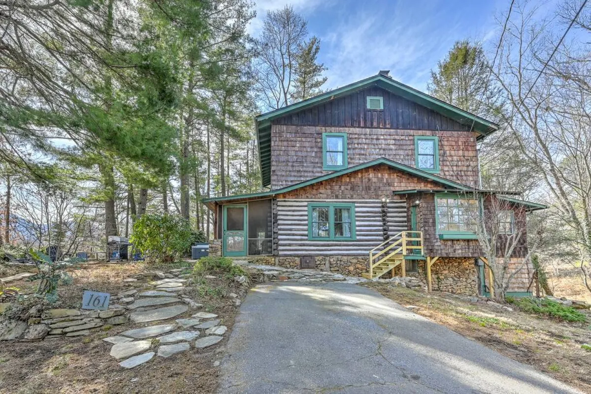 The log cabin exterior, with its green trim and stone pathway, is warmly nestled among trees. The porch offers a view for watching sunsets and provides a welcoming entrance that hints at the proximity to nature trails and areas for outdoor play.