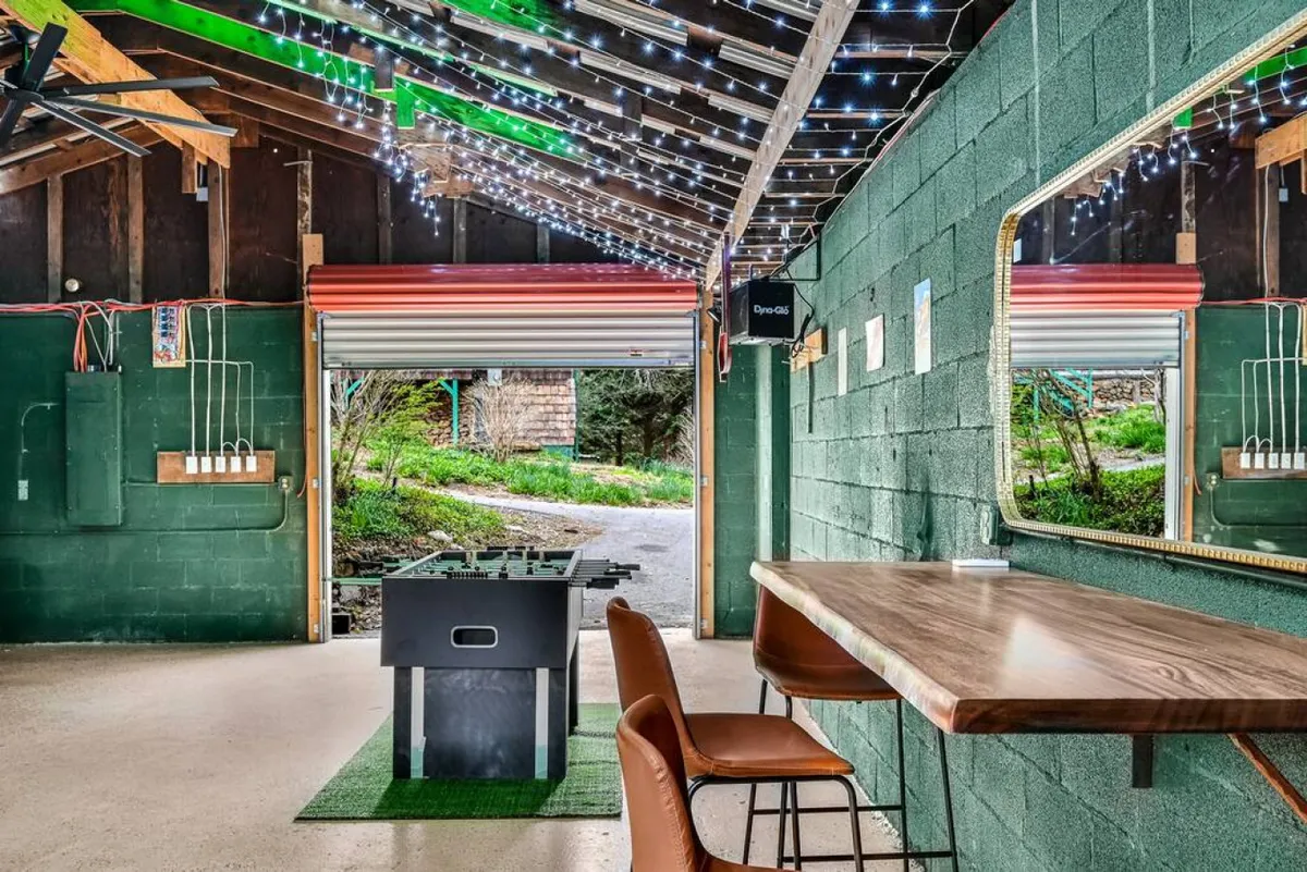 A lively game room with a foosball table as the centerpiece, a bar counter for socializing, string lights overhead, and an open garage door that blends indoor and outdoor spaces. The setup invites friendly competition and relaxation with a touch of festivity.