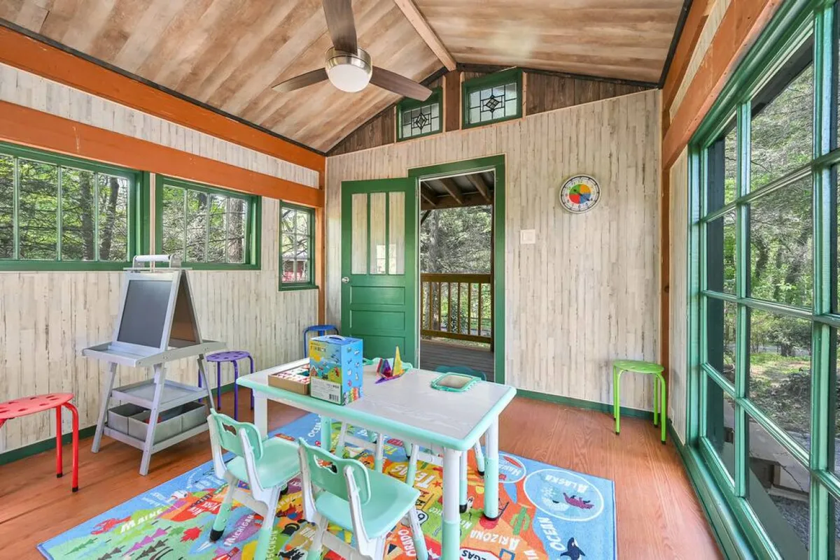 A cheerful and creative indoor play space with a colorful rug, child-sized furniture, vibrant green door, and large windows letting in natural light. The room is clearly designed for children, encouraging imaginative play, arts and crafts, and group activities.