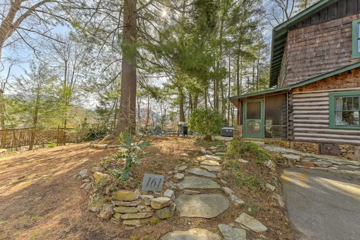 A charming log cabin exterior with stone pathways winding through lush greenery. The green window frames and surrounding trees create an inviting, rustic atmosphere. The porch looks ideal for sipping coffee or relaxing outdoors, while the backyard seems perfect for quiet moments together.